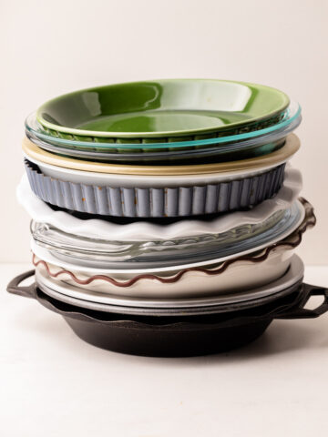 A stack of 14 different pie plates and dishes in assorted materials, including glass, ceramic, and metal