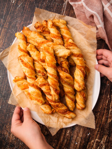 Two hands holding a platter of baked puff pastry cheese twists.