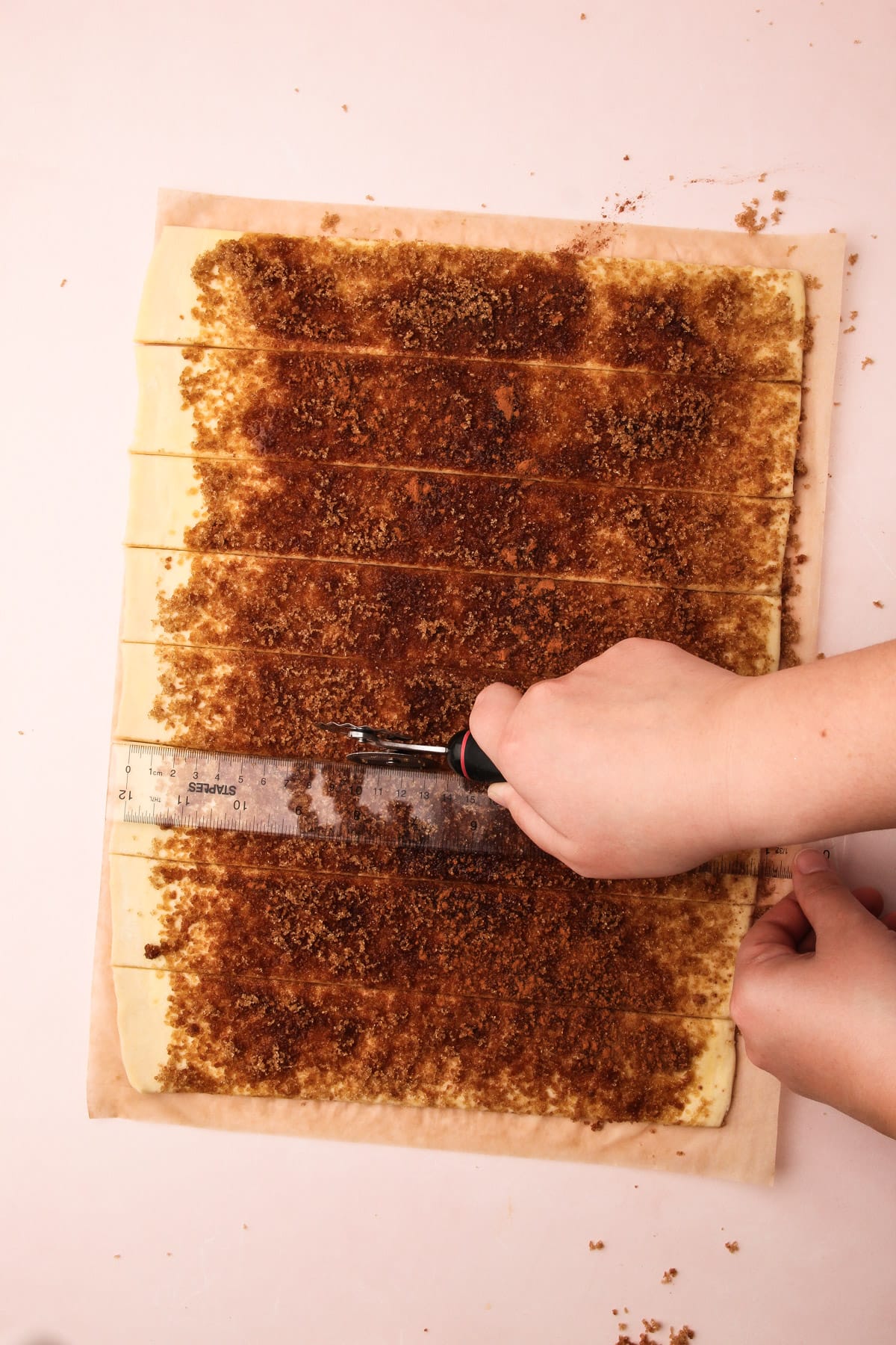 A hand using a pastry wheel and a ruler to cut cinnamon-covered puff pastry into strips for puff pastry cinnamon rolls.