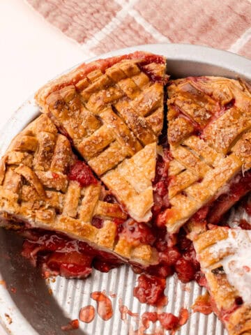 A baked and sliced strawberry rhubarb pie in a pie tin, with bits of the strawberry rhubarb filling leaking out.